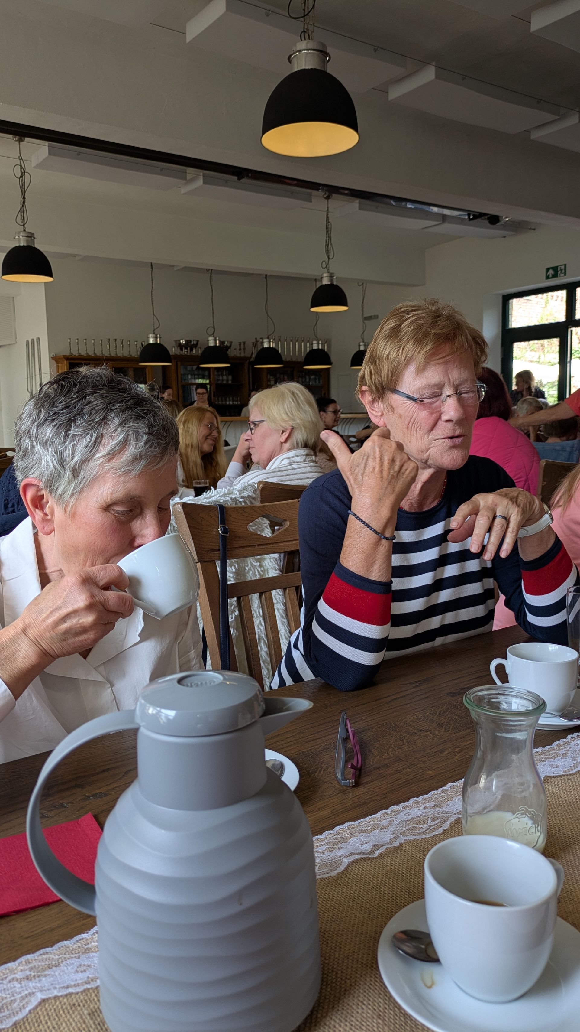Zwei Personen sitzen an einem Café-Tisch mit Tassen und einer Kaffeekanne und unterhalten sich in einem hellen Speisesaal.
