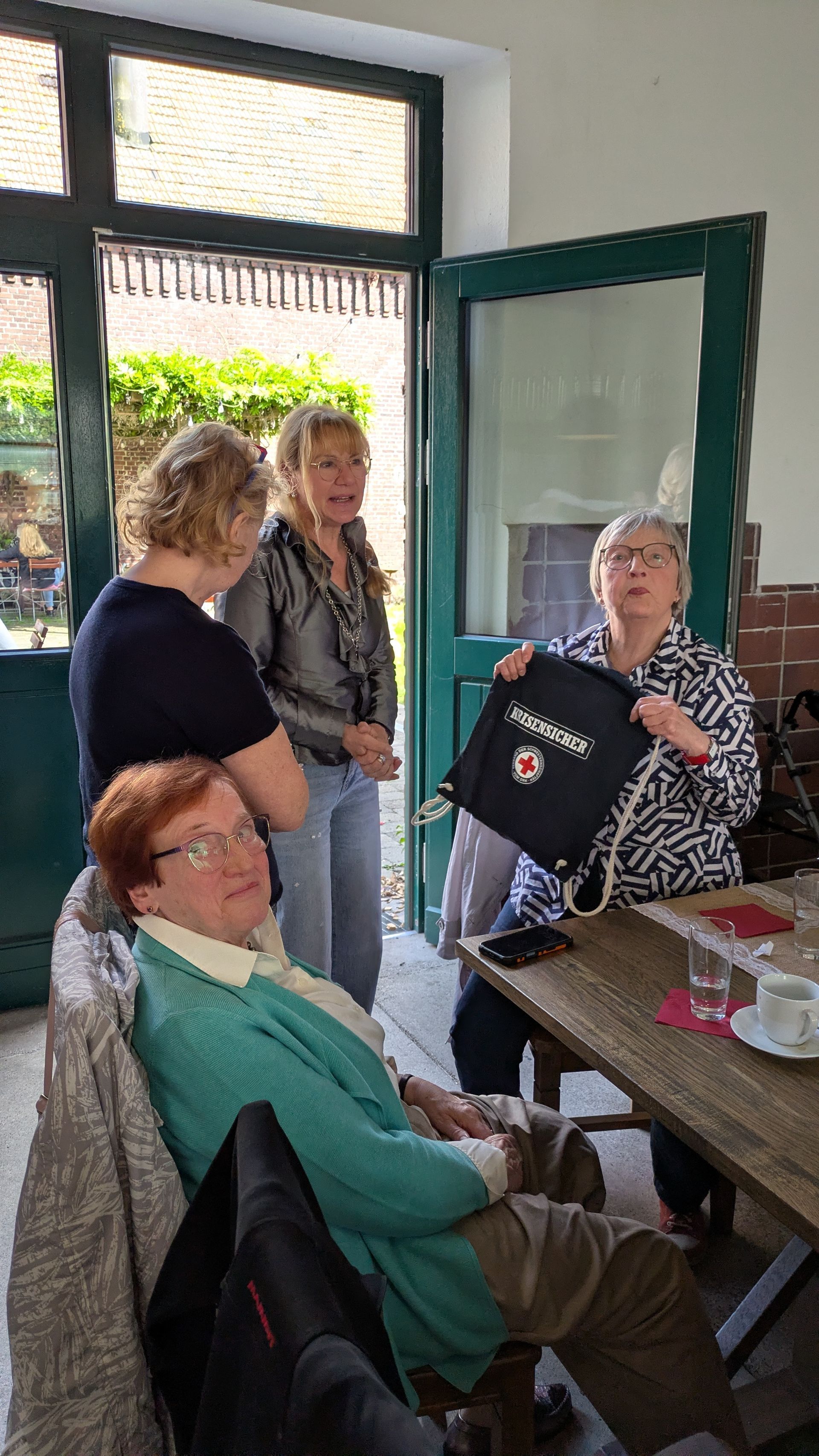 In einem kleinen Café unterhalten sich Menschen in einem Hauseingang, während eine Frau eine schwarze Tragetasche hochhält.