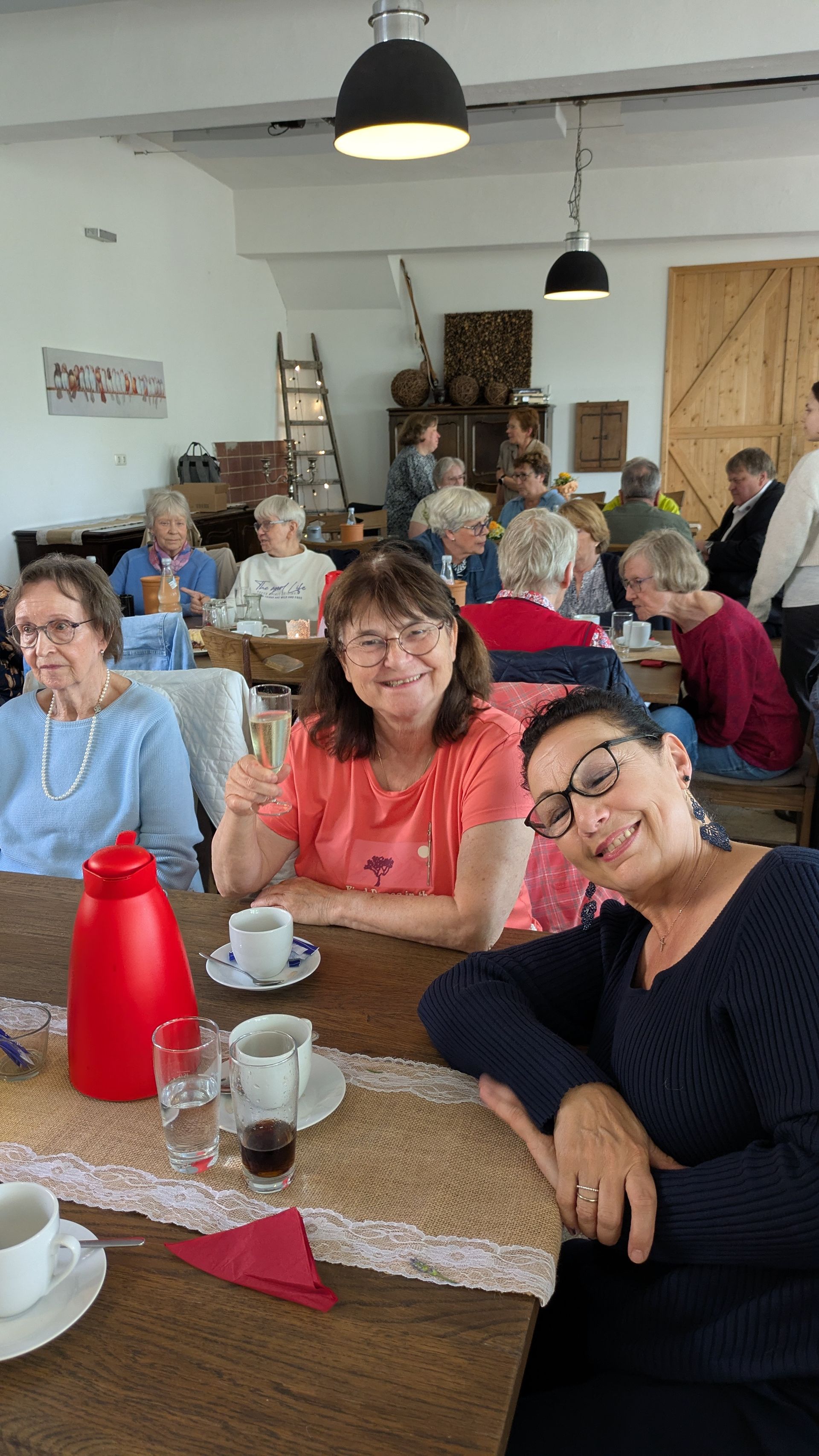 Menschen sitzen an Tischen in einem hellen Café, unterhalten sich und lächeln; vor ihnen stehen Tassen und eine rote Wasserflasche.
