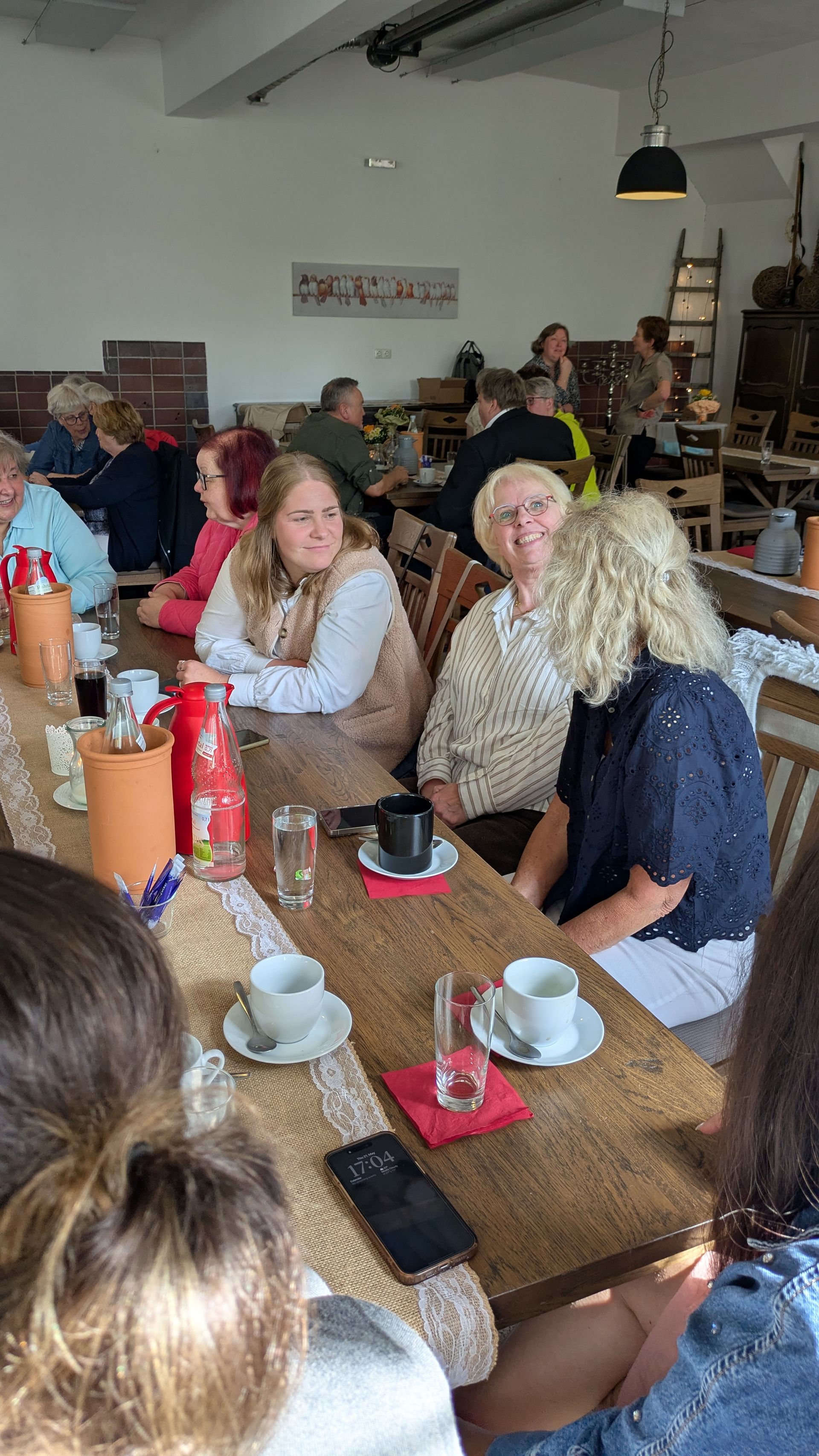 Eine Gruppe sitzt an einem Café-Tisch; im Hintergrund sind Kaffeetassen, eine rote Soßenflasche und eine Leiter zu sehen.