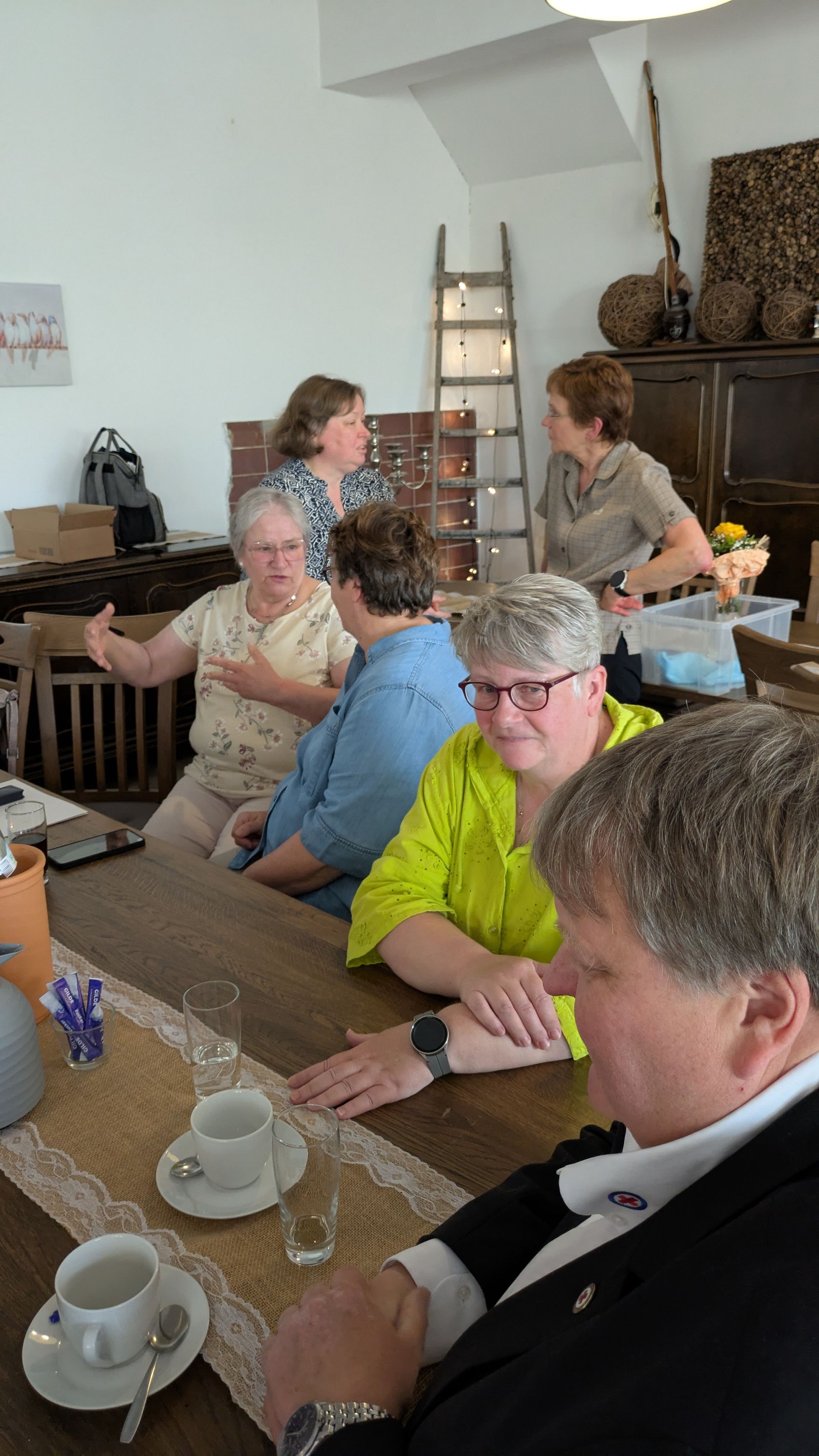 Menschen sitzen an Tischen in einem gemütlichen Café und unterhalten sich bei einer Tasse Kaffee; im Hintergrund sind eine Leiter und ein Kamin zu sehen.