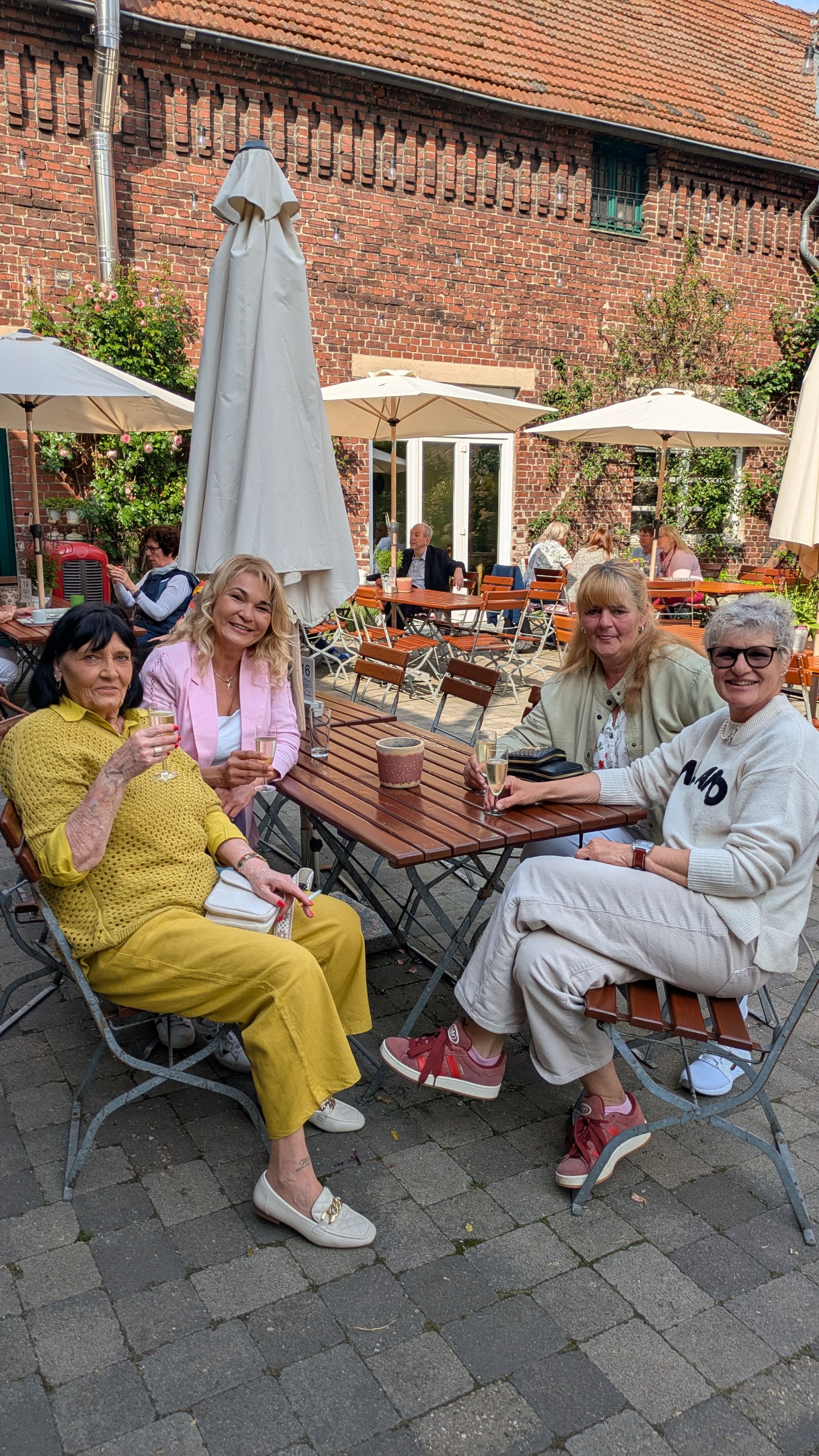 Drei Personen sitzen an einem Tisch in einem Straßencafé unter Sonnenschirmen, im Hintergrund ist ein Backsteingebäude zu sehen.