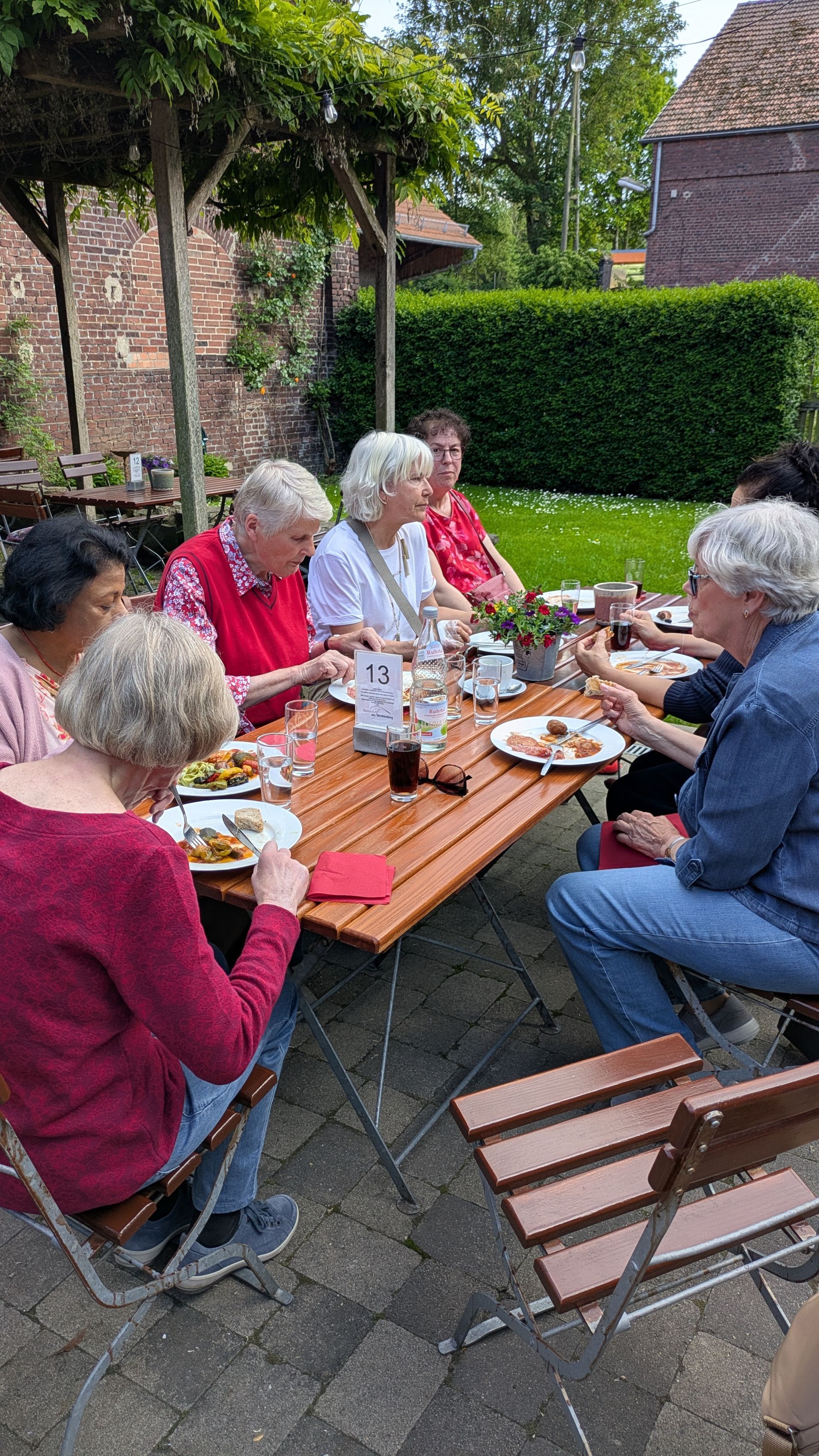 Menschen sitzen im Freien an einem Picknicktisch und teilen Speisen und Getränke in einem von Backsteinmauern umgebenen Innenhof.