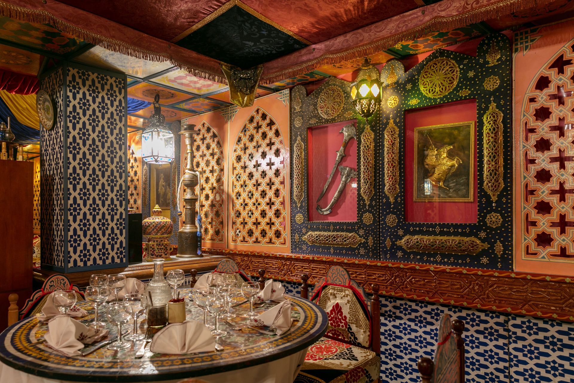 Salle à manger opulente avec murs à motifs, lampes ornementées et table dressée avec argenterie et verrerie.