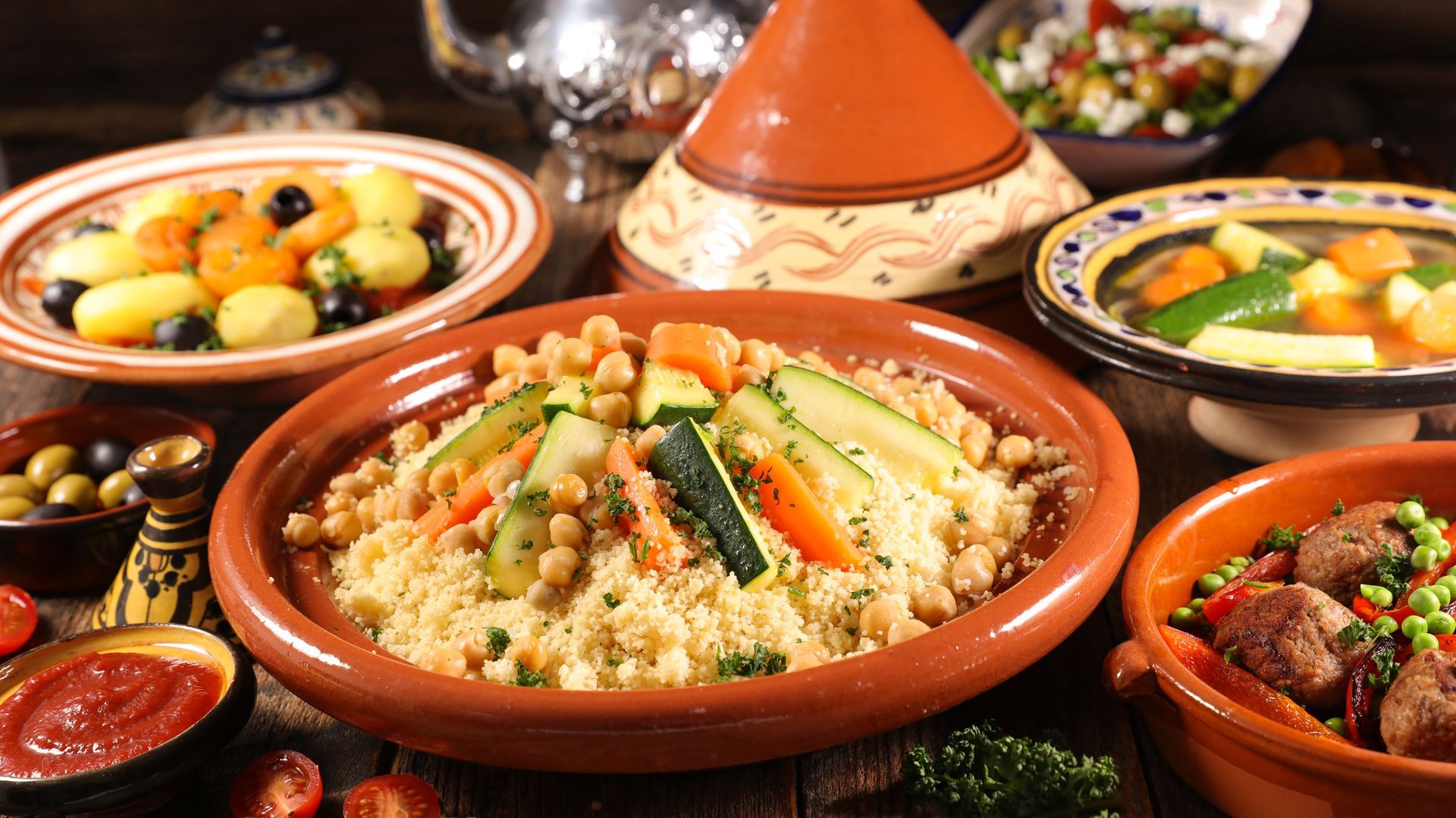 Assortiment de plats marocains présentés dans des tajines et des bols colorés sur une table, avec du couscous au centre.