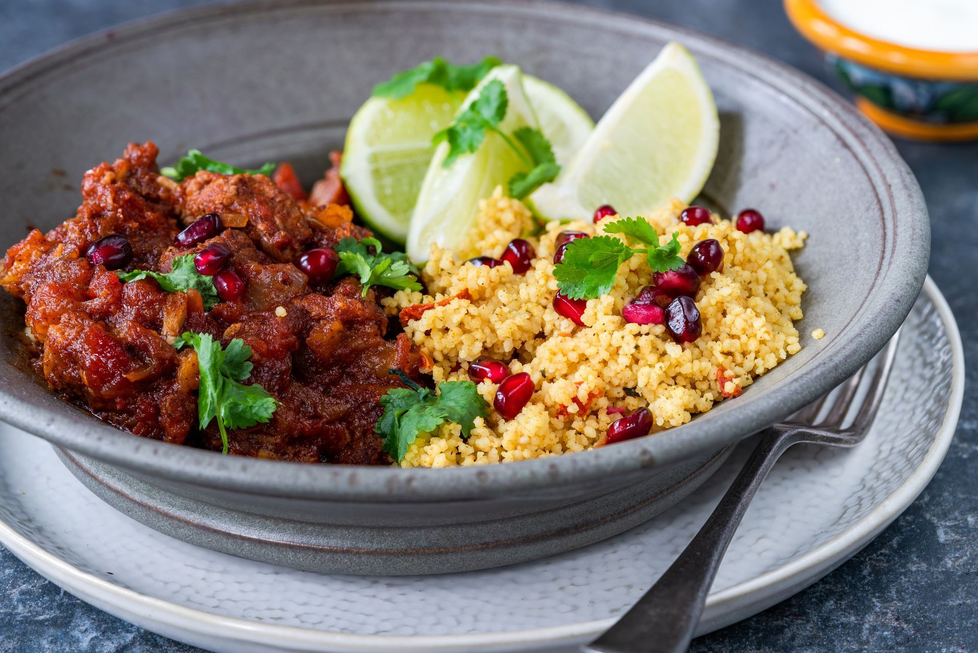 Bol gris avec du couscous et de la viande épicée garnie d'herbes, de quartiers de citron vert et de graines de grenade
