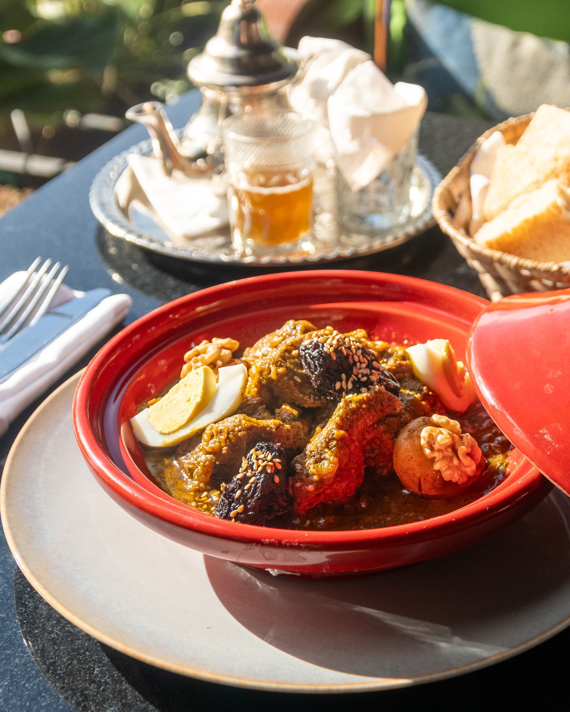 Un bol rouge de tajine aux pommes de terre sur une table de café ensoleillée, avec du thé et du pain en arrière-plan.