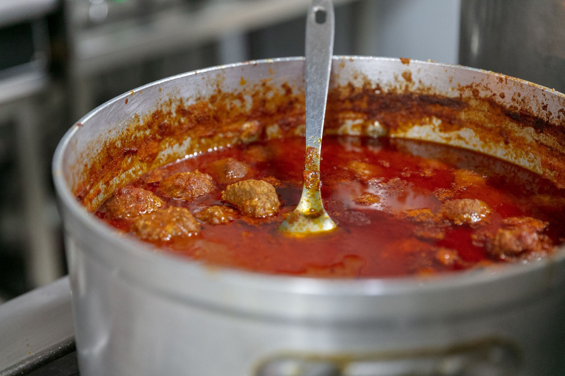 Grande marmite en métal contenant un ragoût ou une sauce rouge, avec une louche, laissant apparaître des morceaux de viande qui mijotent à l'intérieur.