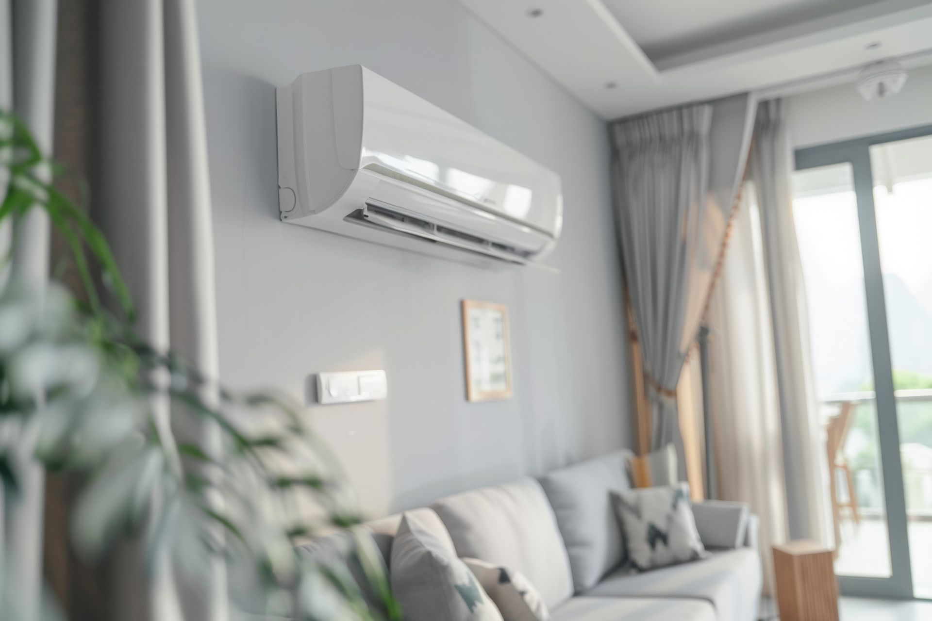 Salon lumineux avec climatiseur mural, canapé et fenêtre ensoleillée avec rideaux.