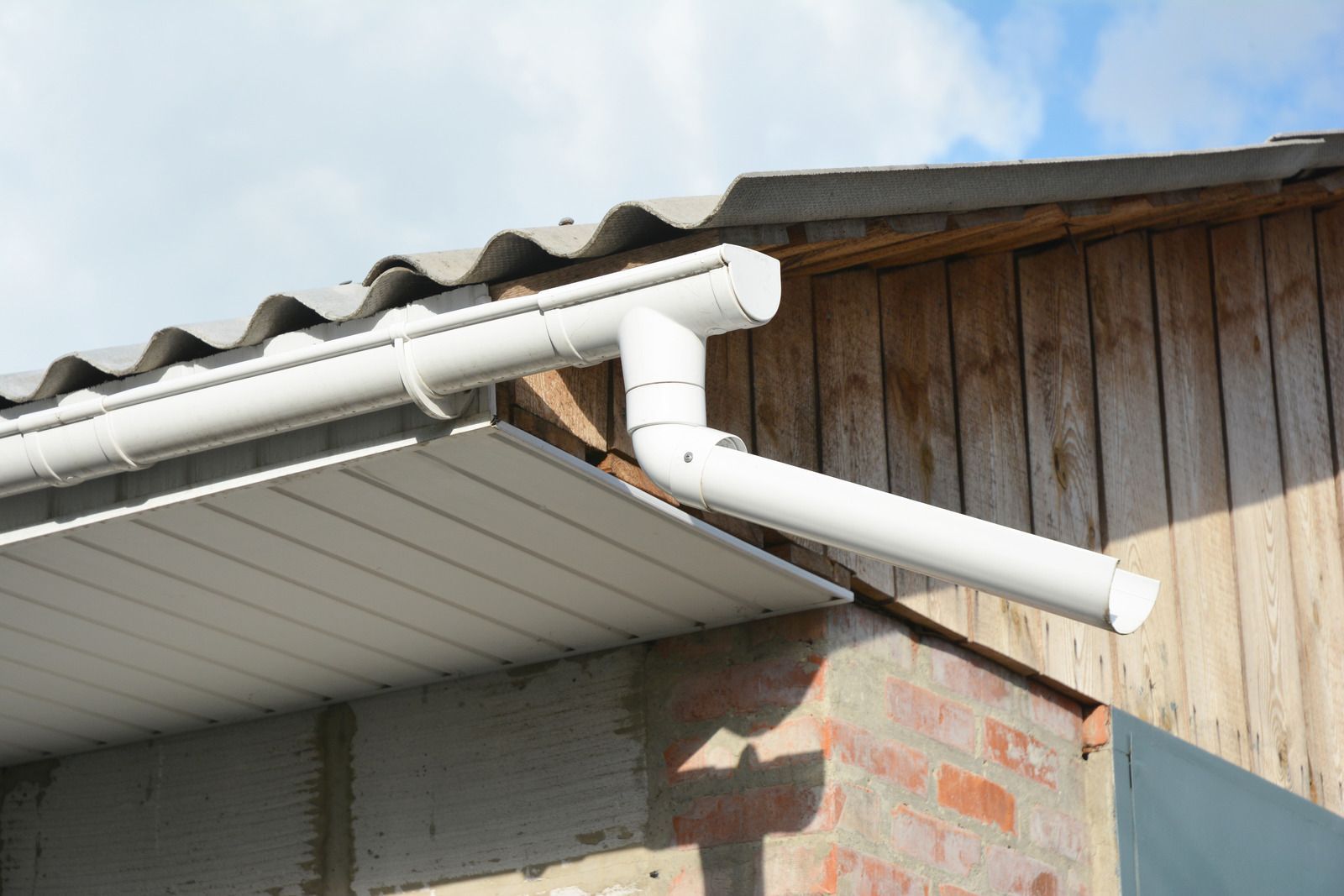 White gutter and downspout attached to a corrugated roof beside a wooden wall
