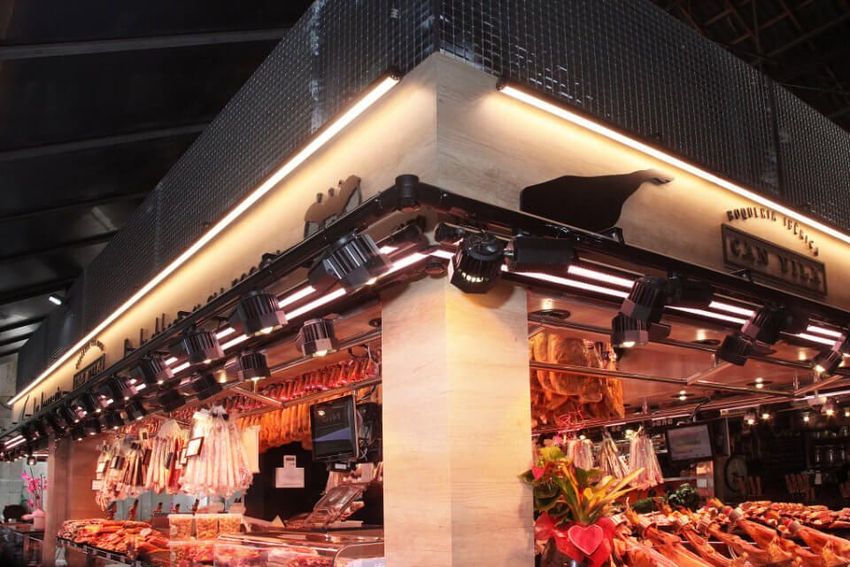Rincón de un puesto de mercado cubierto muy iluminado, con carnes y productos colgando bajo una luz cálida.