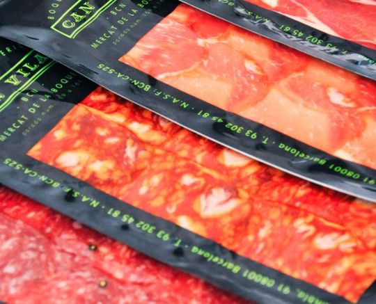 Surtido de embutidos envasados ​​en bandejas negras con etiquetas verdes y contenido rojo anaranjado.