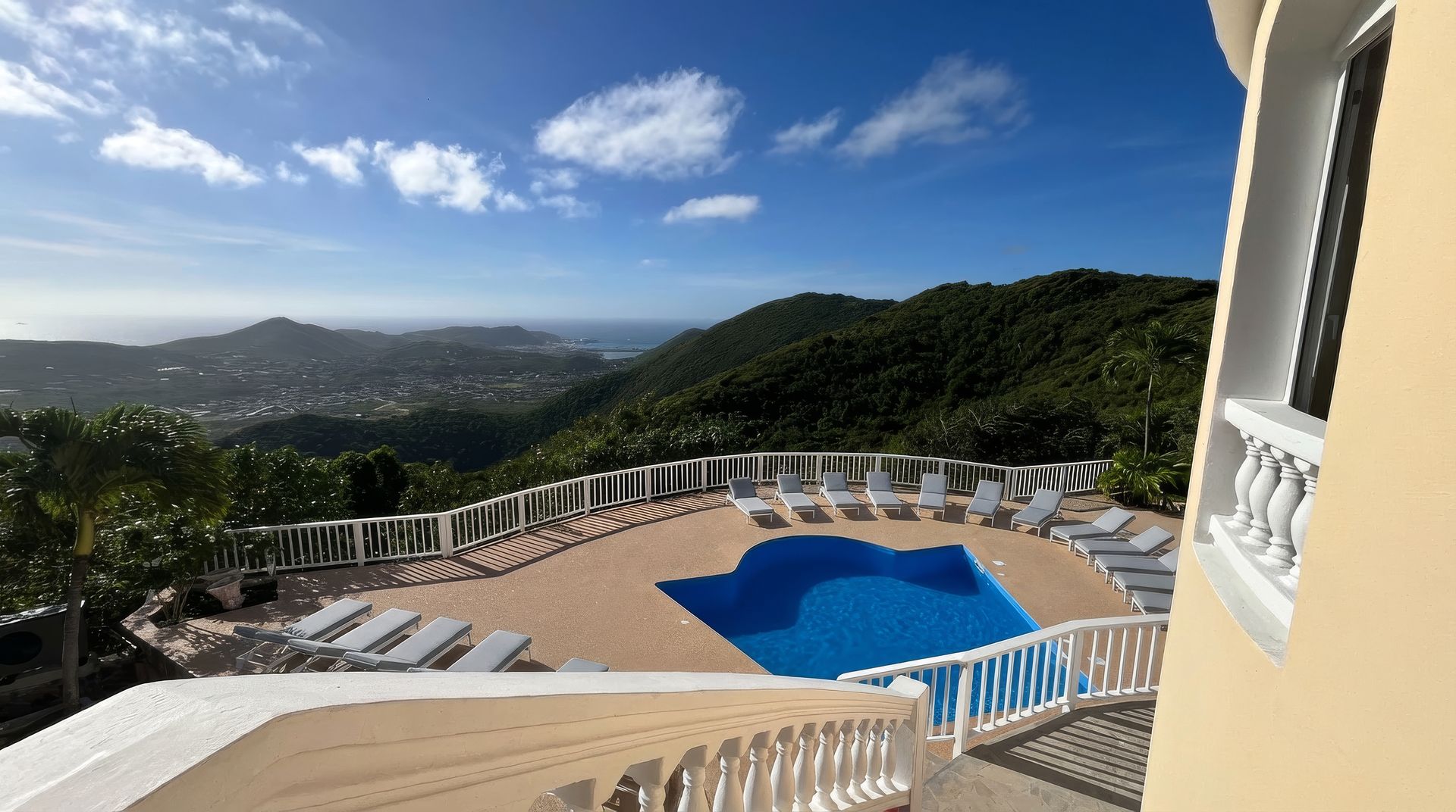 Swimming pool overlooking a lush mountain landscape and distant town; sunny sky.