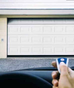 Una persona en un automóvil usa un control remoto para abrir una puerta de garaje blanca.