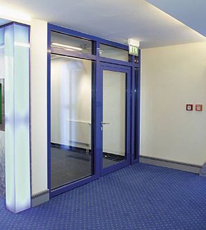 Puerta de vidrio con marco azul y paneles laterales en el pasillo de un edificio. Alfombra azul y paredes blancas.