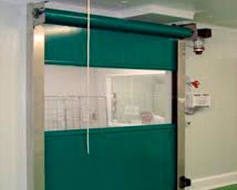 Puerta enrollable rápida verde con ventana, instalada en una habitación con paredes blancas. Se ve una persona tras la ventana.