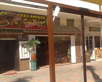 Exterior del restaurante Tres Amigos con toldo marrón, ventanales y fachada de ladrillo. Una planta en maceta se encuentra junto a la puerta principal.