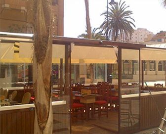 Zona de restaurante al aire libre con paredes de cristal y toldo retráctil. Mesas y sillas visibles en el interior.