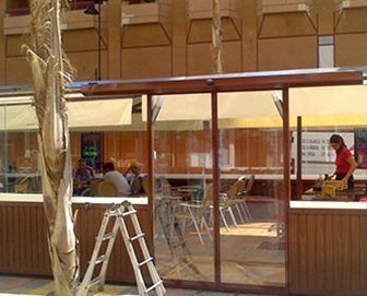 Una cafetería con paneles de vidrio transparente y marcos de madera. Una persona trabaja en un mostrador. Los clientes se sientan en mesas en el interior. Cerca hay una escalera.
