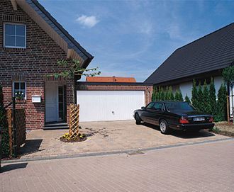 Casa de ladrillo con puerta de garaje blanca y un coche negro aparcado en la entrada. Día soleado.