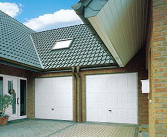 Puertas de garaje dobles blancas con pared de ladrillo adyacente, techo de tejas grises y ventana pequeña.