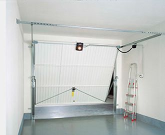 Puerta de garaje parcialmente abierta, revelando un interior con escalera. Puerta blanca, marco de metal, suelo gris claro y paredes blancas.