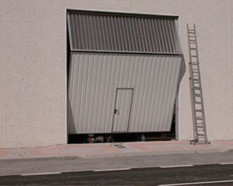 Una gran puerta industrial blanca, parcialmente abierta en el exterior de un edificio, con una pequeña puerta en su interior. Una escalera se encuentra a la derecha.