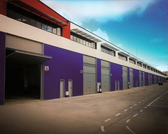 Largo almacén con exterior morado y blanco, puertas grises y un techo rojo bajo un cielo azul.