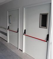 Dos puertas blancas con ventanas rectangulares y barras de salida rojas en un pasillo.
