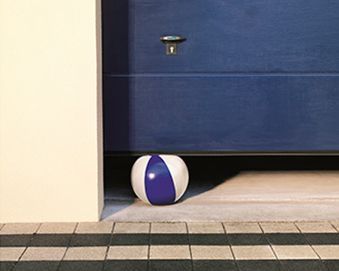 Pelota de playa bajo una puerta de garaje azul entreabierta. A la izquierda hay una pared blanca y en primer plano un suelo de baldosas.