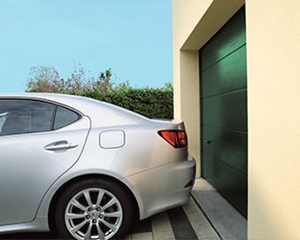 Coche plateado aparcado cerca de una puerta de garaje verde oscuro. Un entorno soleado con cielo azul y un exterior de color canela claro.