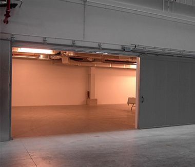 Abra una puerta corrediza de metal hacia una habitación luminosa y vacía con paredes blancas y piso de concreto.