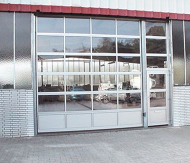 Gran puerta de garaje industrial con paneles de vidrio transparente que reflejan los coches en el interior. Tiene una pequeña puerta a la derecha.