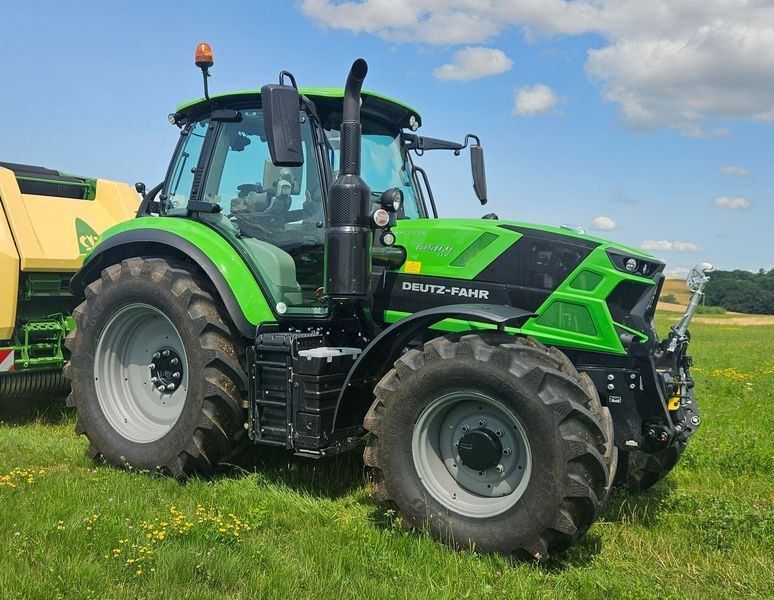 Un tracteur Deutz-Fahr vert dans un champ, tirant une presse à balles de foin sous un ciel bleu.