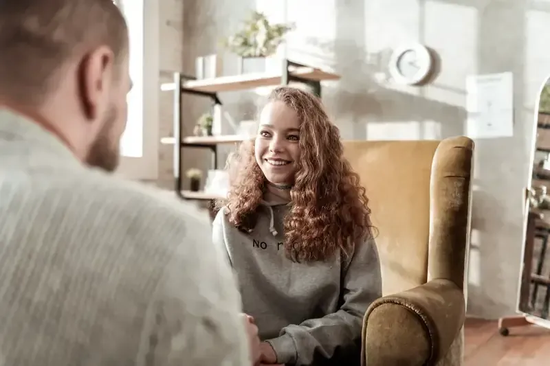 Persona sonriendo, sentada en una silla, frente a otra. Interior con luz tenue.