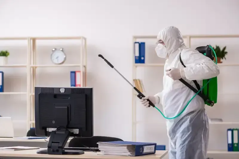 Persona con traje de materiales peligrosos rociando desinfectante en una oficina.