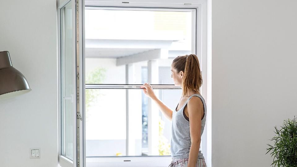 Eine Person zieht in einem weissen Zimmer ein Insektenschutzrollo an einem offenen Fenster herunter.