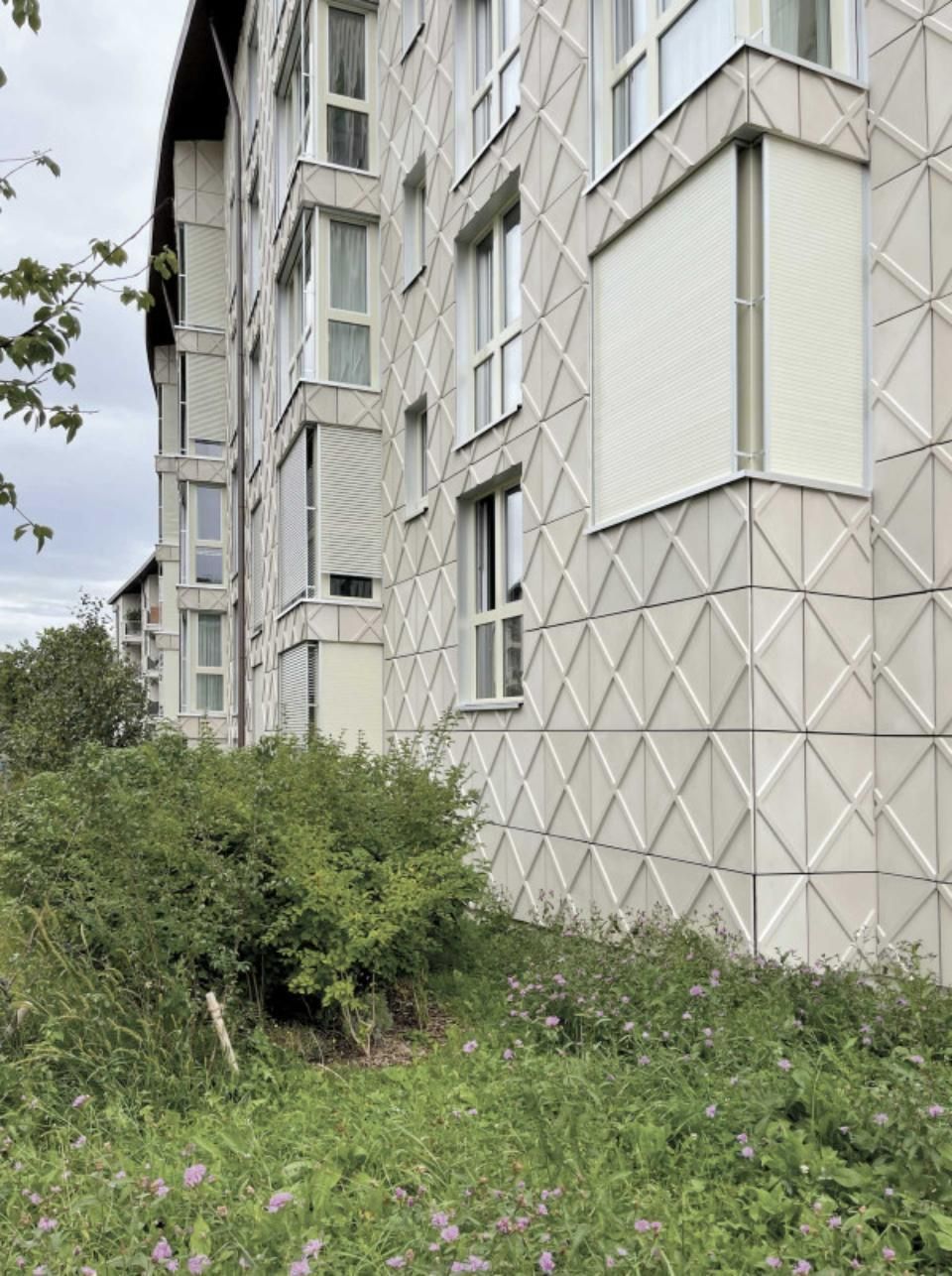 Eine weisse Wohnhausfassade mit einem geometrischen Rautenmuster, die teilweise von grünen Sträuchern verdeckt wird.