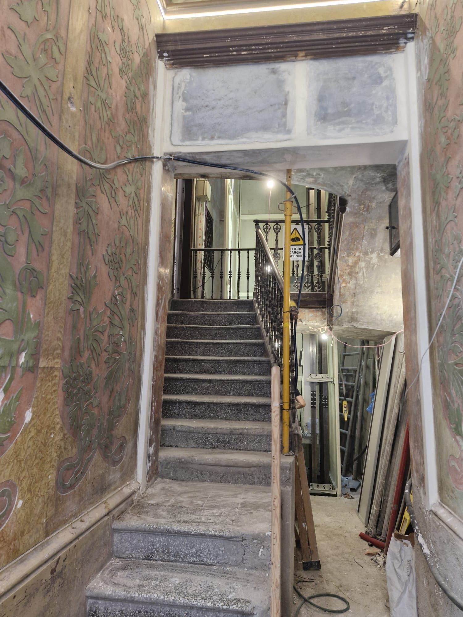 Escalera que asciende con paredes envejecidas y pintura decorativa. Puerta oscura en la parte superior.