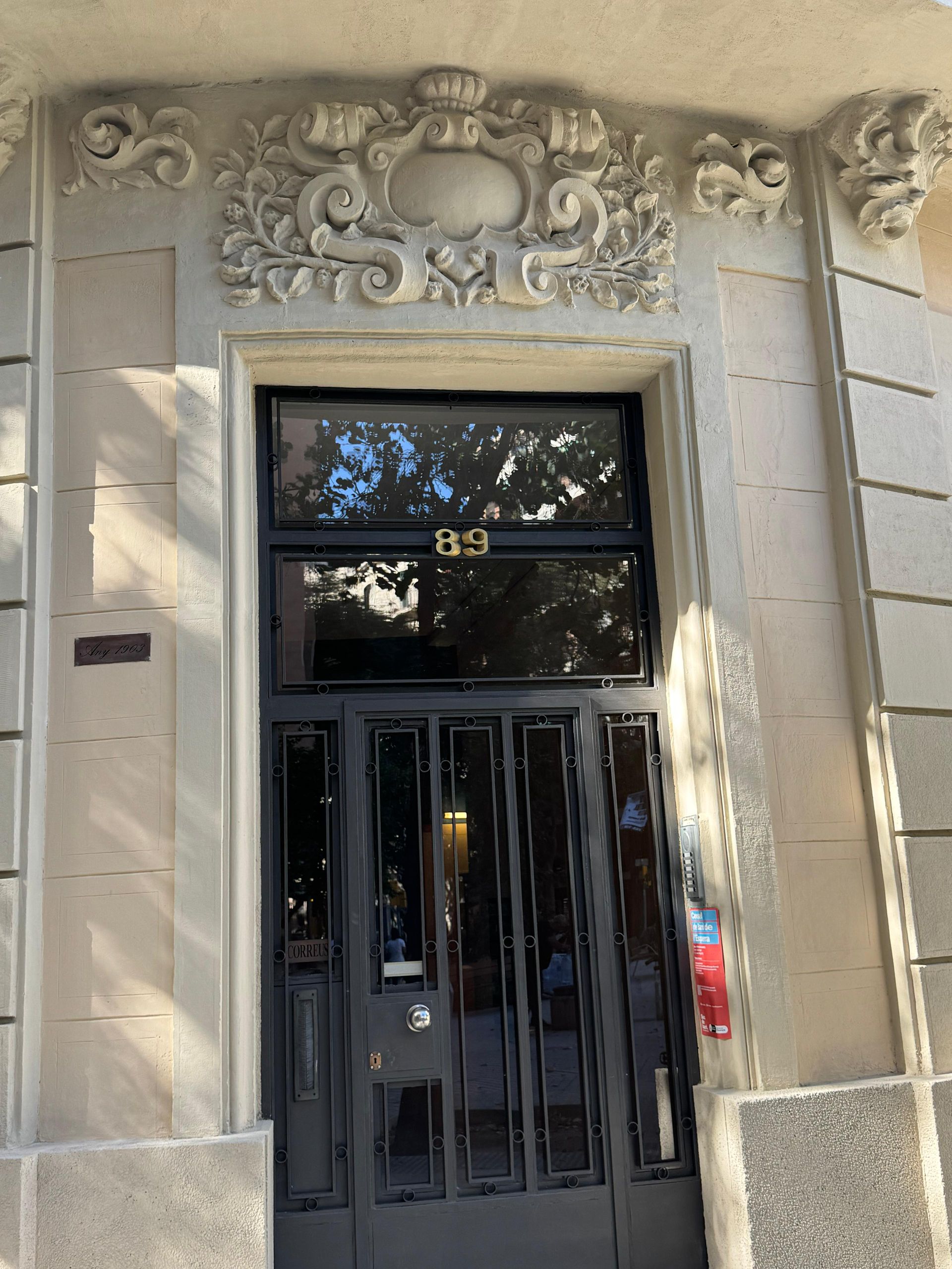 Puerta gris con paneles de vidrio y ornamentación de piedra en la parte superior, edificio número 89.