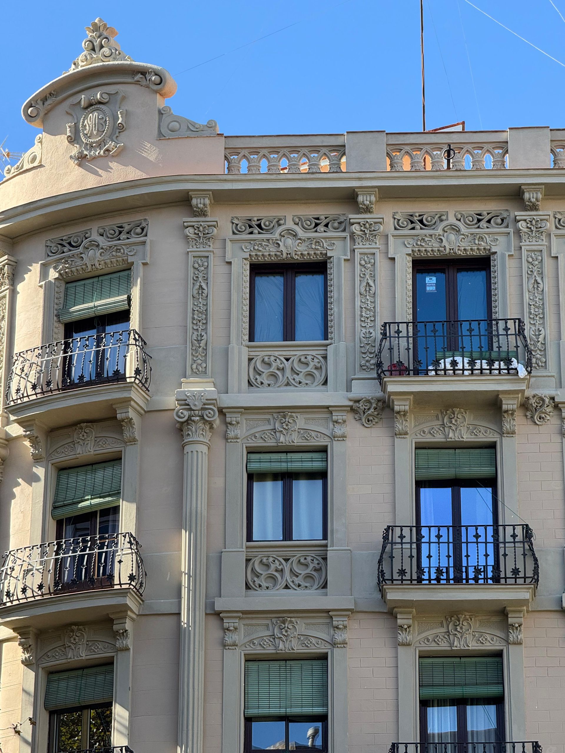 Edificio de apartamentos color beige con tallas ornamentales, balcones y persianas, contrastando con un cielo azul despejado.