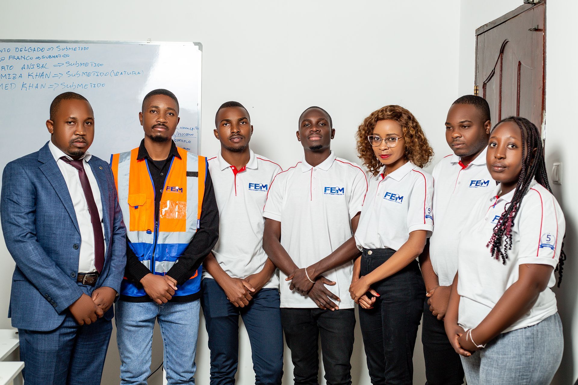 Group of seven people posing in an office, wearing various attire.