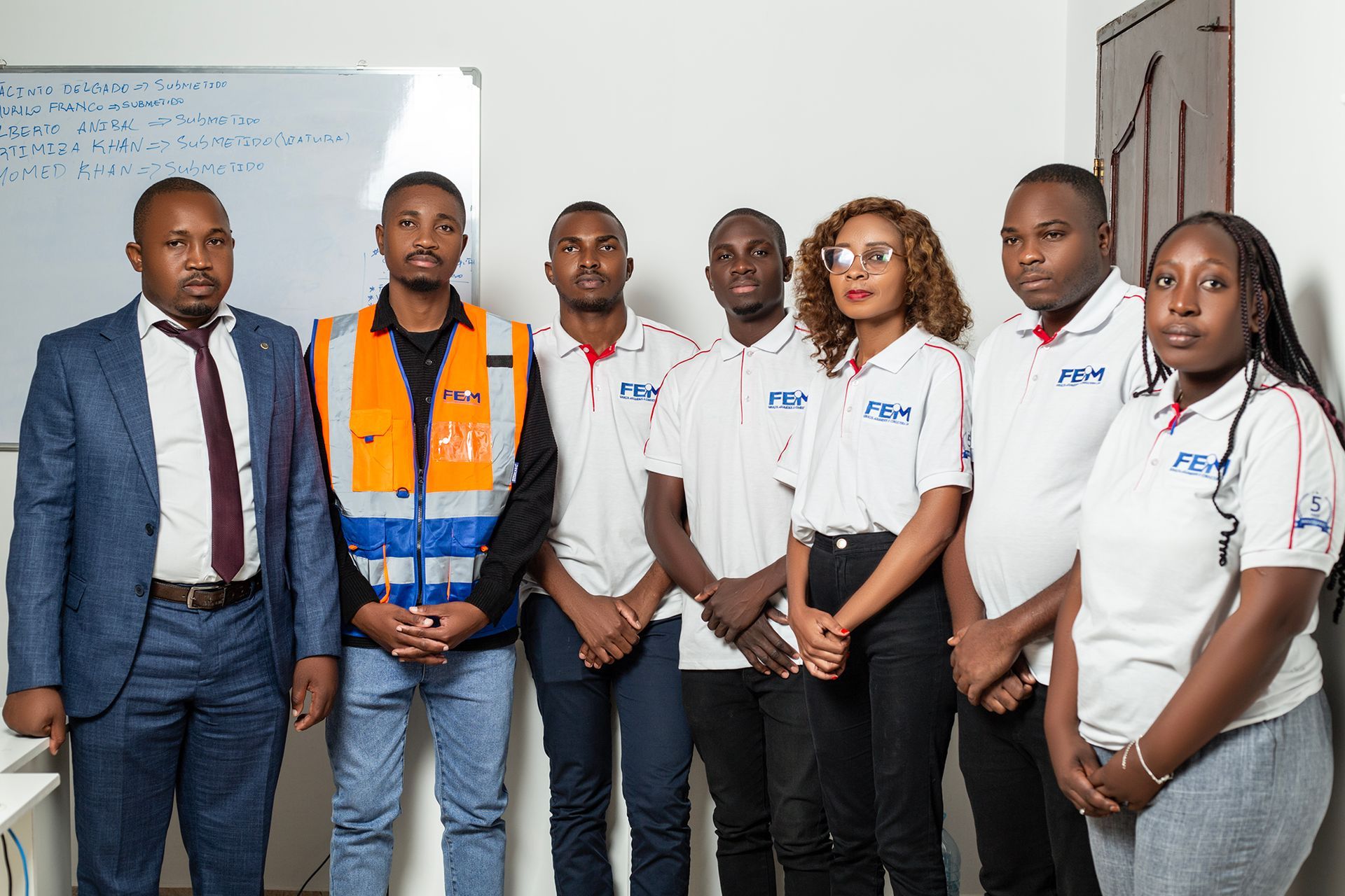 Group of seven people standing in a room; some wearing company uniforms and one in safety vest.