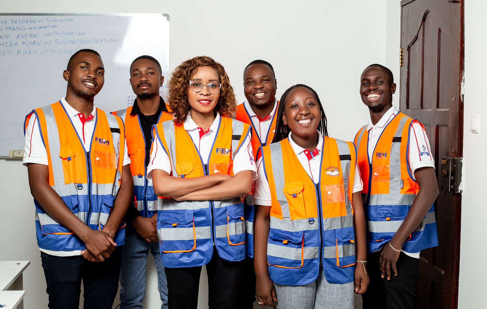 Group of six people wearing orange and blue safety vests, posing indoors.