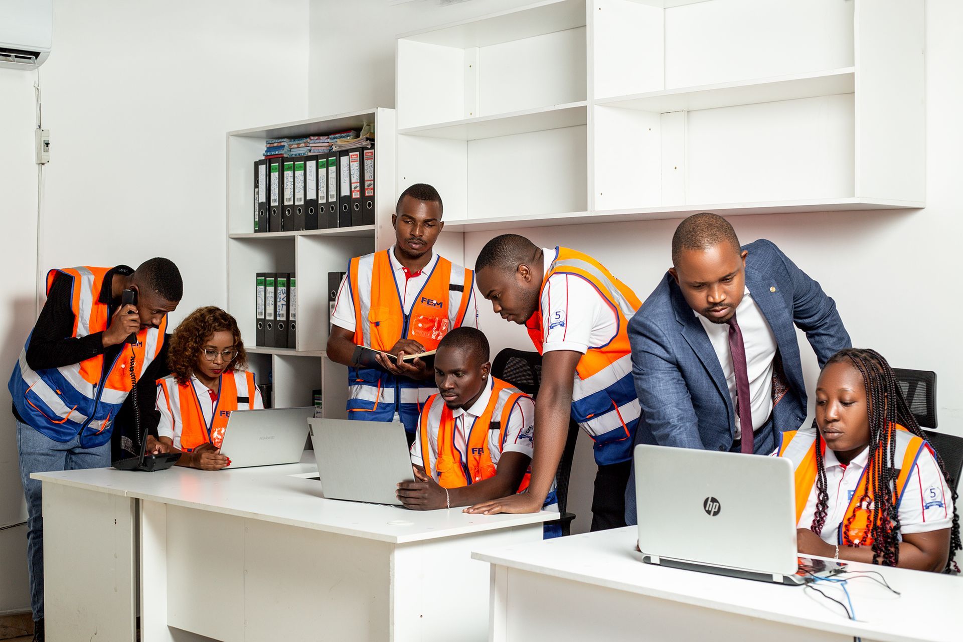 People in orange vests and business attire working in an office with laptops.