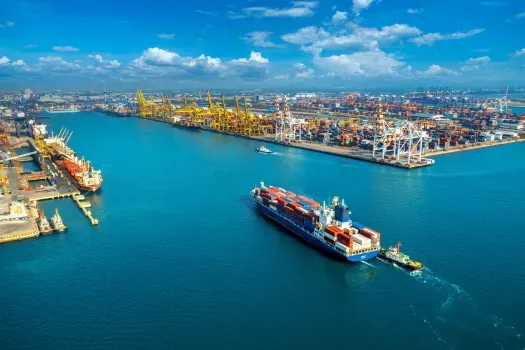 Aerial view of a busy blue harbor with cargo ships, cranes, and a container port skyline