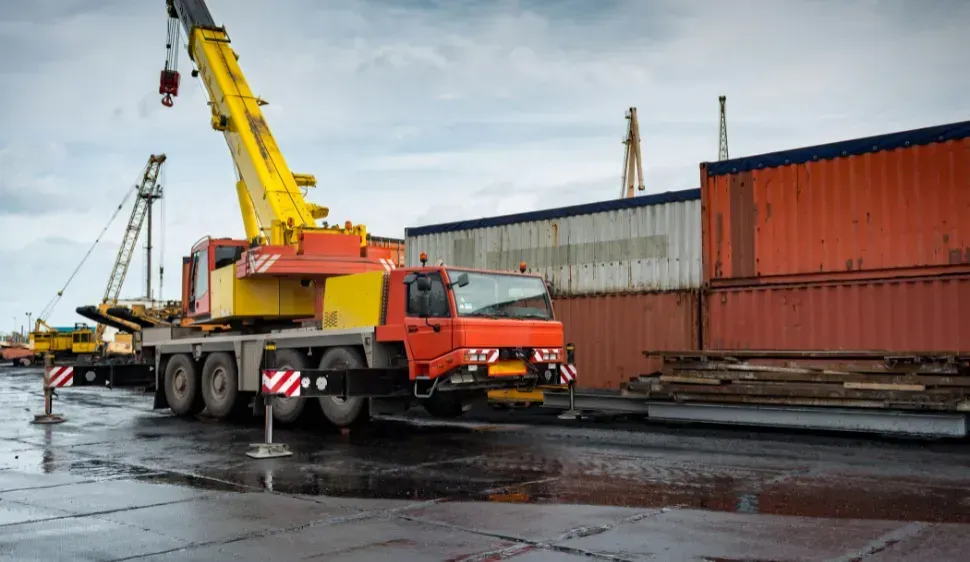 Mobile crane lifting, orange and yellow, near shipping containers at a dock. Cloudy sky.