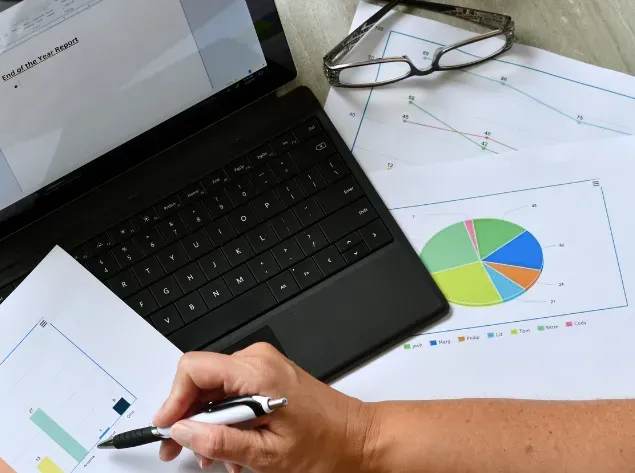 Hand writing notes beside a laptop, eyeglasses, and colorful charts on a desk