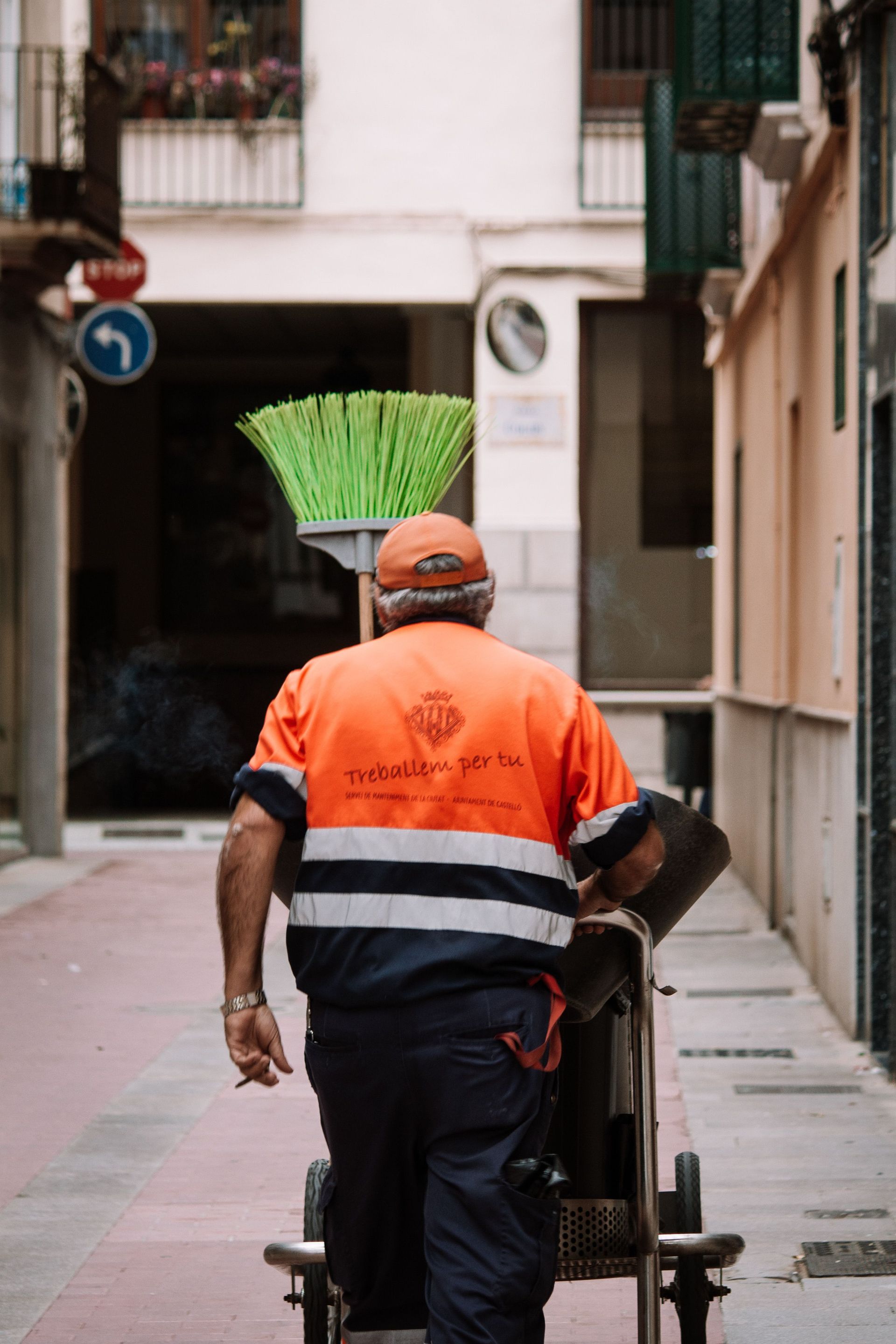 Trabajador con uniforme naranja empujando un carrito de basura por una calle estrecha.