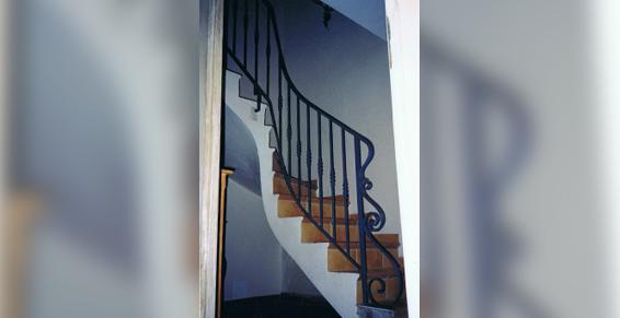 Escalier avec marches en bois et rampe en fer forgé noir incurvée vers le haut dans un espace aux murs blancs.